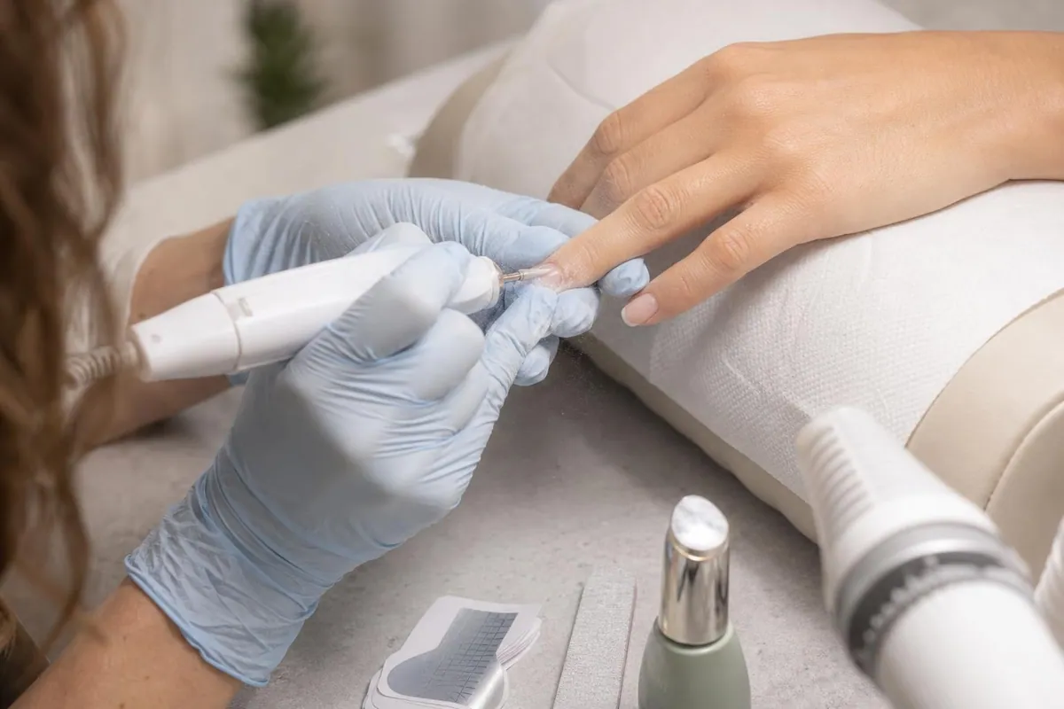 Gros plan sur les mains gantées d’une technicienne ponçant la repousse d’un ongle en gel sur la main d’une cliente